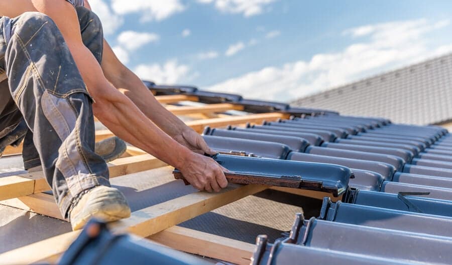 A worker from Titan Exteriors is applying blue roofing tiles on a roof, showcasing skilled roofing installation. A worker from Titan Exteriors is applying blue roofing tiles on a roof, showcasing skilled roofing installation.