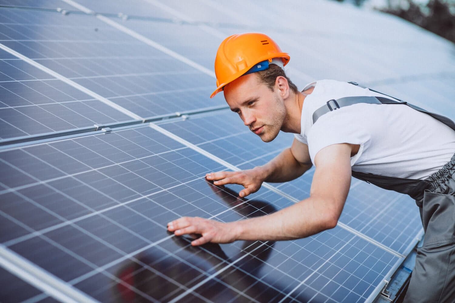 A worker in overalls and a hard hat is installing solar panels for Titan Exteriors on a sunny day. A worker in overalls and a hard hat is installing solar panels for Titan Exteriors on a sunny day.